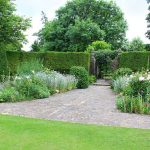 Photo showing a formal garden with a neatly clipped yew tree hedge (taxus baccata). In the centre of the hedge is an iron gate, while the foreground is softened with herbaceous plants, roses, evergreen shrubs and bushes. Crazy paving forms the pathway, which is edged by a recently mown green lawn of fine grass.